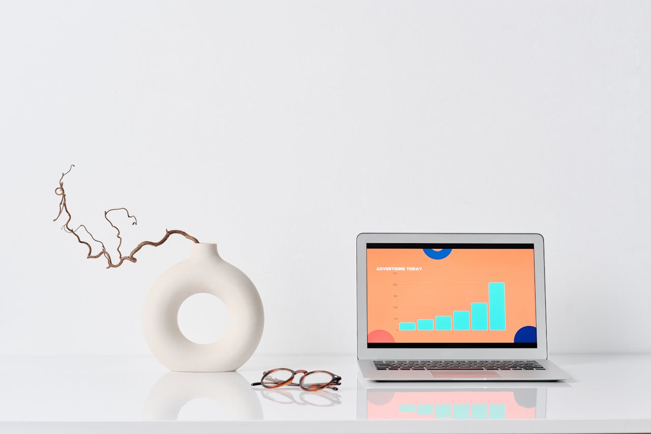 A minimalist workspace with a laptop, ceramic vase, and eyeglasses on a white desk.